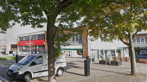 A pedestrianised square with the Post Office top left, with three other shop fronts to the right of the Post Office. A couple of trees are at the front of the square with two black bins and a van behind the tree on the left.