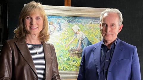 Presenters Fiona Bruce and Philip Mould standing in front of the brightly-coloured artwork which depicts women in a rural landscape. Bruce is wearing a brown jacket and striped top and Mould is wearing a blue suit jacket and shirt