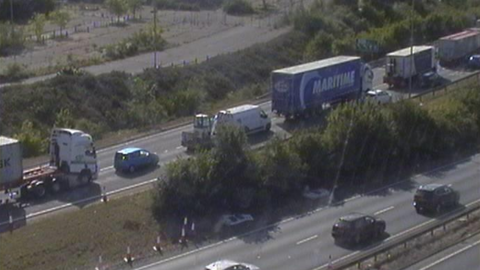Traffic camera image showing queueing traffic including cars and lorries on the offside lane of the two lanes on the left of the hedge-lined central reservation. An open space with some vehicles parked in it is visible on the left of the road, possibly the site of a demolished factory.