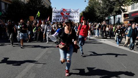 A woman with a megaphone runs towards the camera as the crowd approaches behind her