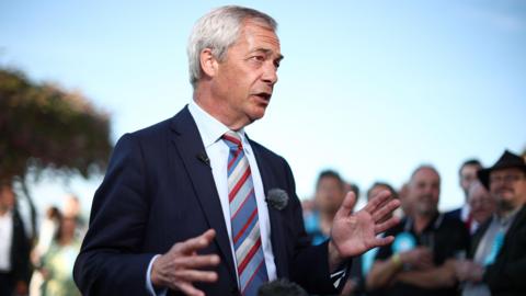 Reform UK leader Nigel Farage attends a post-election event at the Staffordshire County Showground. He is wearing a suit and tie and gesturing with his hands and there are people gathered around him, listening.