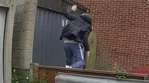 A man in tracksuit bottoms and a padded jacket is seen on the other side of a wooden fence, running away.