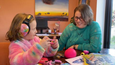 A young girl in a pink fluffy jumper with ear defenders on her head plays with a toy at a table while her mother in a green jumper watches on.