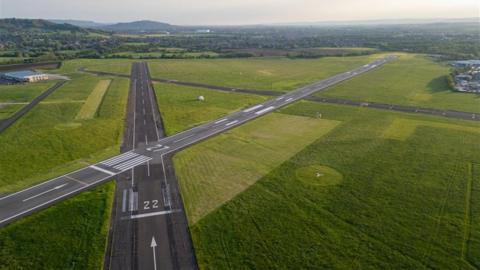 The runways at Gloucestershire Airport intersecting one another, surrounded by grass. The Golden Valley can be seen in the background on what looks like a summers evening.