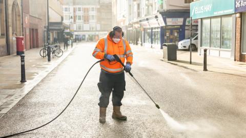 A man in an orange vest with a mouthguard and ear protectors on is jet washing a street.