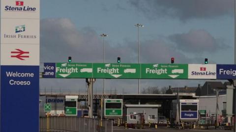 Three green signs above different lanes directing freight traffic on to Irish Ferries service to Dublin are seen at the Port of Holyhead in Holyhead. To the left of these is a large blue and white 'Welcome' sign with the various transport companies listed. There are no vehicles driving towards the barriers.