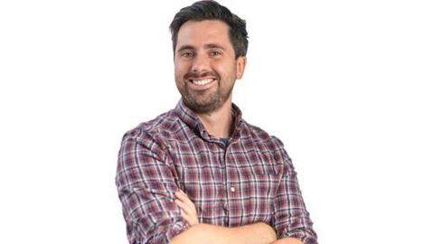 A promotional shot of Jack Murley, smiling with arms crossed and wearing a check shirt. He has short dark hair and dark stubble.