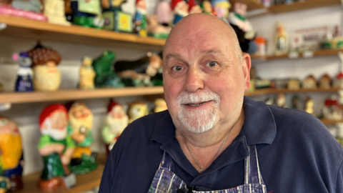 Alistair Dent, a former butcher turned gnome doctor. Dent is standing inside his former butcher shop in Hilgay. Behind him is rows and rows of different gnomes. Dent is looking directly at the camera and is wearing a blue polo shirt with a striped blue and white apron over the top.