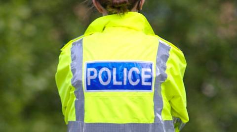 A female police officer stands facing away from the camera with her fluorescent yellow hi-vis jacket bearing the word 'police' on a large blue badge, with trees out of focus in the background.
