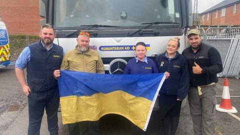 Volunteers holding a Ukrainian flag in front of a silver humanitarian aid lorry