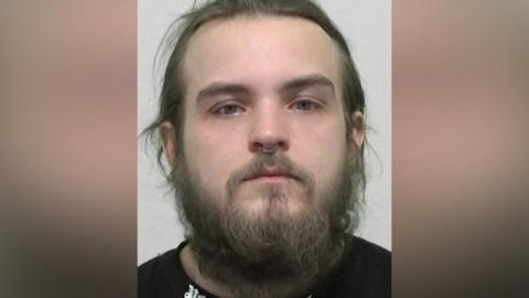 Mugshot of Kennedy. He has a straggly beard and long messy brown hair, with a ring through the septum of his nose.