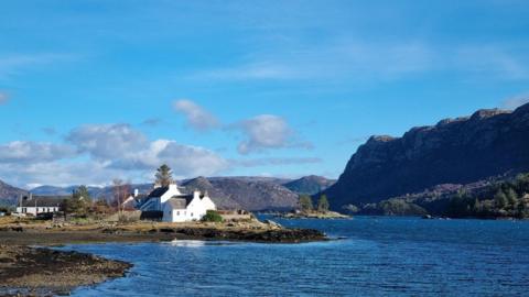 Bright blue sky over a bright blue sea inlet with mountains to the right and the sun bouncing off a white houses in the centre