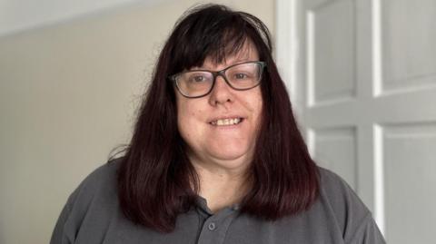 Middle aged woman wearing a grey polo shirt smiling at the camera. She has dark brown shoulder length hair with a fringe and is wearing black framed glasses.