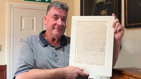 A man holding a letter signed by Mary Queen of Scots. He is wearing a short sleeved light blue shirt. There is a closed door in the background and pictures on the wall.