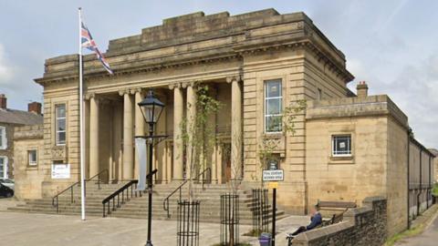 Mercer Hall is a large stone-built property with six large pillars at the entrance, above steps with black railings. It has a flagged frontage with a Union Jack flag flying from a flagpole and a vintage street lamp.
