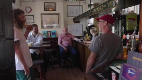 Several people inside a pub talking and drinking, with beer glasses on tables and at the bar, and wall pictures and signs behind them.
