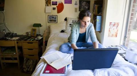 Young woman sat cross-legged on a bed using a laptop with an open book in front of her to the right