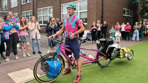 Luke Grenfell Shaw returning to Bristol after completing the challenge on his pink-and-blue tandem bicycle. He is wearing a pink and blue cycling top and navy shorts.
