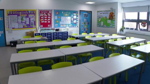 An empty classroom. There are rows of seats which are green in colour. There are signs on the boards around the classrooms of children's work.