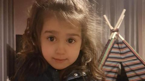 Sara Shariff aged four. She's is smiling at the camera and is standing in front of a stripey wigwam tent in a house