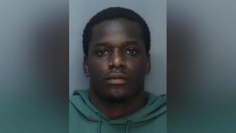 A man wearing a green hooded jumper stares directly at the camera