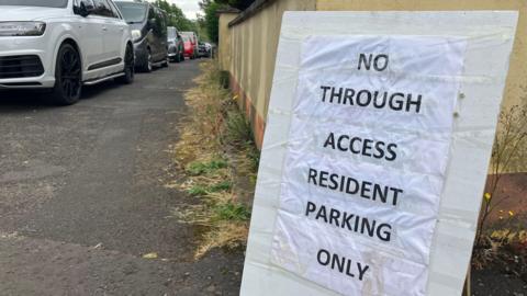 A sign rests against the wall. It stands in front of a number of parked cars and reads: No through access, resident parking only