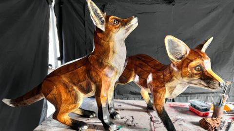 Two 3D fox lanterns. The lanterns are placed on a table top. One of the foxes is looking up, while the other is designed to look like it is walking along.
