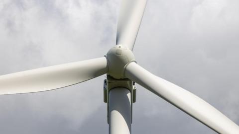 A close up of a wind turbine's hub and three blades. The machine is white in colour and is pictured against a cloudy, grey sky.