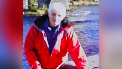 Bryan Perry, with white hair wearing a red coat and a blue fleece, standing in front of a river
