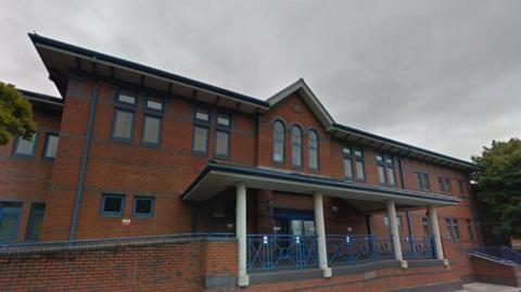 A red-brick court building which has ramp access to the left hand side and step access to the right hand side. There are blue railings, and trees either side of the building.