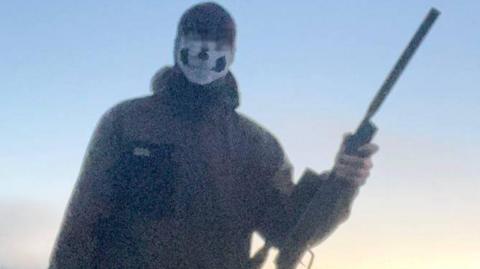 A blurry image of the boy standing in a field and holding a gun. He is wearing a mask with a skull design on it. His eyes above are blurred. He is wearing a dark waterproof jacket. 