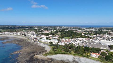 A grey, rocky beach. The sea is in front of it. In the background are houses and green fields. The sky is blue. 