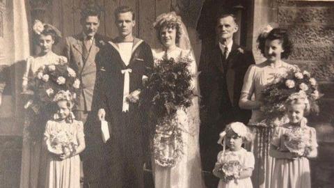 A black and white photo of a group of people at a wedding outside a church, including the bridge and groom.