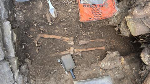 A partially excavated human skeleton lies exposed in a shallow soil pit, with leg and arm bones clearly visible.
Archaeological tools, including small brushes and a plastic scoop, are scattered around the dig area.
Surrounding the site are stones, tree roots, and a bag.
