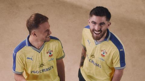 Leo Scienza and Ryan Manning are both wearing the new yellow and blue commemorative shirts. They're both laughing. Leo looks over at Ryan while Ryan grins up towards the camera. The shirts feature a P&O Cruises logo and the words '1976 FA Cup winners'. 