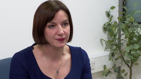 A woman with mid-length dark hair, a silver necklace and a blue top sits on a chair next to a plant.