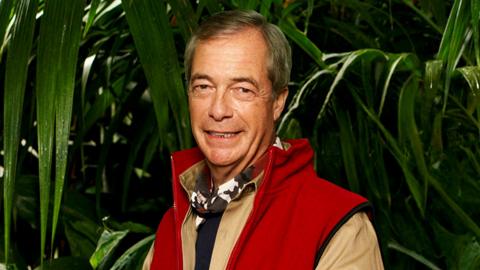 A man with short grey hair stands in front of a jungle background of dense foliage, made up of long, green leaves that hang down around him. He's standing with his hands clasped together, and wears a khaki (light brown) shirt with a red waistcoat/body warmer over the top. He's got a neckerchief on with a black, white and burgundy camouflage pattern.
