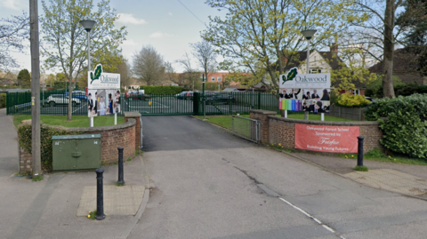 A Google Street image of a school entrance. There are white signs either side of a driveway with a dark green gate at the end. The signs have a green image of a leaf on them and the text "Oakwood School".