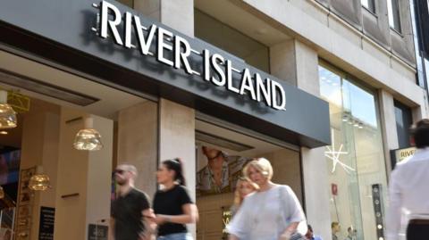 Shoppers walk past a River Island store on Oxford Street in London. It's a sunny day. Two friends are walking side by side and a woman with short blonde hair is walking.