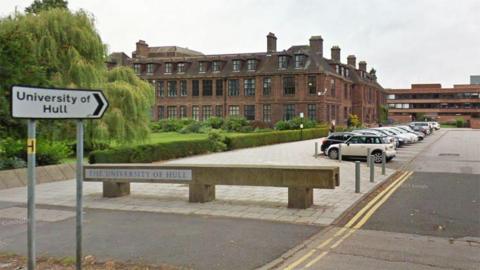 A view of the University of Hull campus from the road with a concrete name sign and cars parked outside the red-brick main building. There are also green trees and hedges.