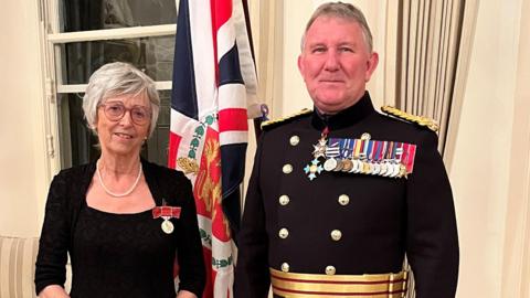 Marguerite Talmage is standing with Lt Gen Richard Cripwell. She is wearing her medal pinned onto a black dress and a string of pearls. He is in full uniform which is pinned with a long row of medals. Behind them is a flag.