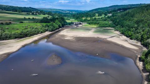 A reservoir, pictured from above against the backdrop of rolling hills and trees. The water is noticeably shallow and much of the bed is dry.