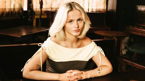 Mel Raeburn, a woman with long blonde hair and wearing a white and black top, sits in a bar with her hands clasped together on the table.