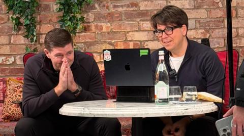 Steven Gerrard and Michael McIntyre are sitting at a table in Emily's restaurant. There are both looking at a Mac book and there is a bottle of water and two glasses on the table. 