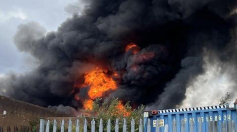 An explosion of orange flames rises into a billowing plume of black smoke above the steel fencing and brown brickwork of the industrial estate.