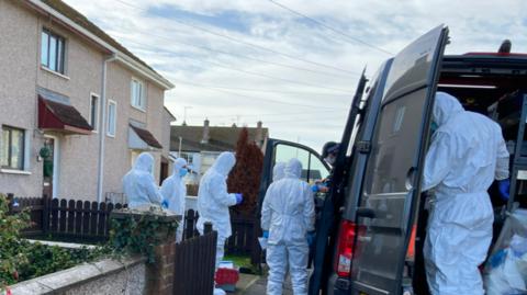 A row of terrace houses with a group of people wearing white overalls standing in front of it. There is a black van parked in front of the houses and the back door is open, with a person standing at the back.
