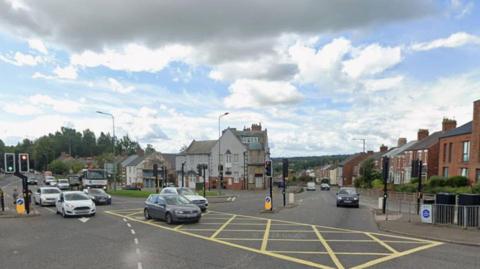 Several vehicles approach a yellow box junction where there are also traffic lights. Residential and business properties are on either side of the road.