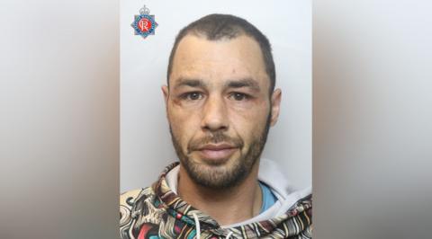 Mugshot of Andrew Alveranga, who has short cropped dark hair and beard. He wears a multicoloured hooded top.