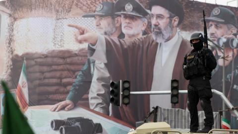 A member of the Iranian security forces stands guard in front of a billboard depicting the new Supreme Leader, Mojtaba Khamenei, and Iranian military and Revolutionary Guards commanders, during a rally to mark Quds Day at a square in Tehran, Iran (13 March 2026)