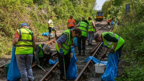 About a dozen male and female volunteers with his visibility vests and litter pickers are collecting waste from a rail line and placing it in blue refuse bags. The rails are surrounded by green shrubs. The sun is shining and a yellow train can be seen in the distance. 